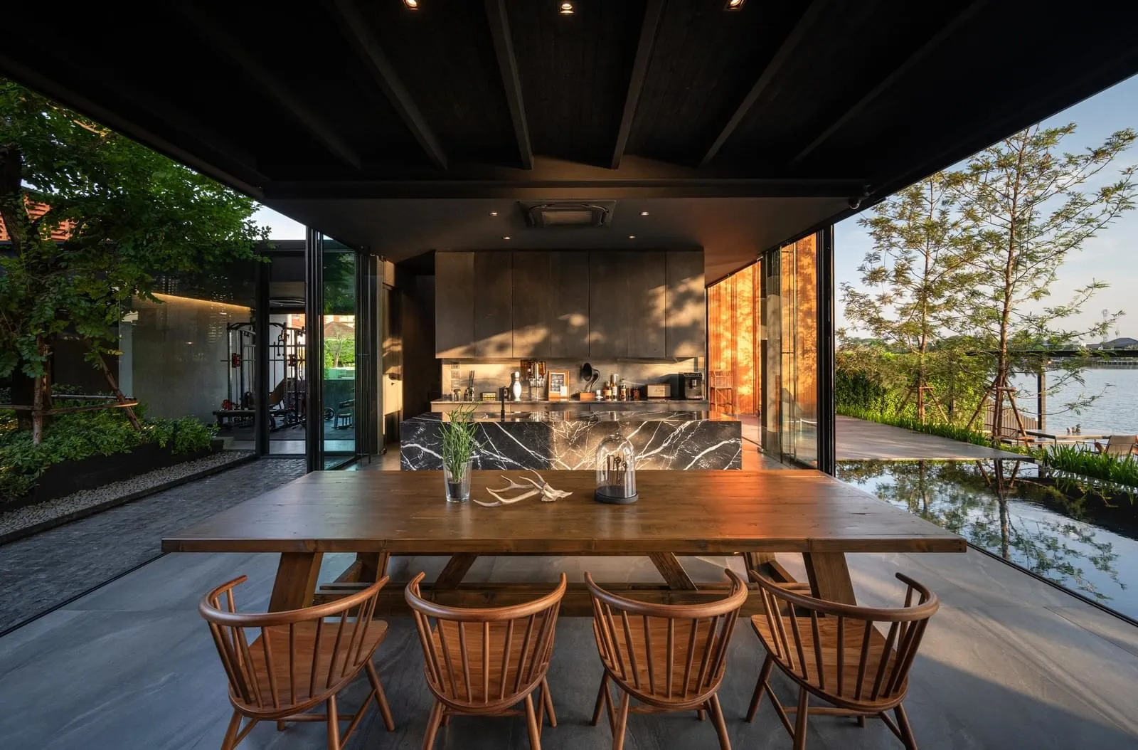 Modern industrial dining room with wooden furniture, marble walls, and glass windows overlooking a serene water feature.