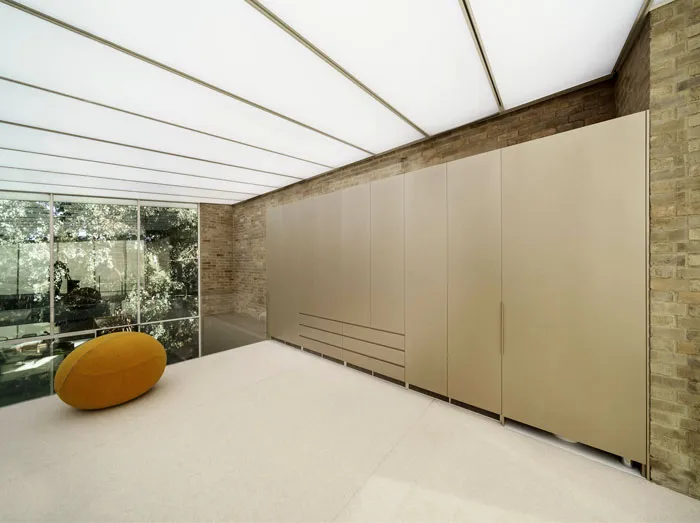 Modern minimalist residential interior with concrete walls and floor, featuring a yellow pouf and large glass windows.