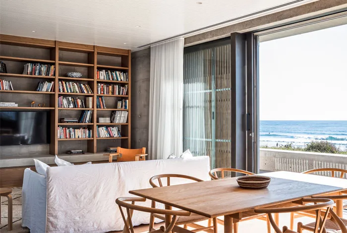 A minimalist living room with a white couch, wooden table, and bookshelf filled with books and decorative items. The room ove