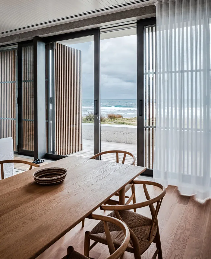 A minimalistic dining room with a wooden table, chairs, and a view of the ocean through large glass doors.