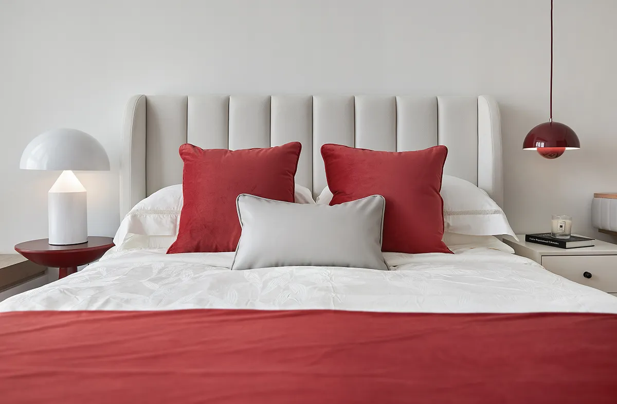 A minimalist bedroom with a white upholstered bed, red and white accent pillows, and a white bedspread.
