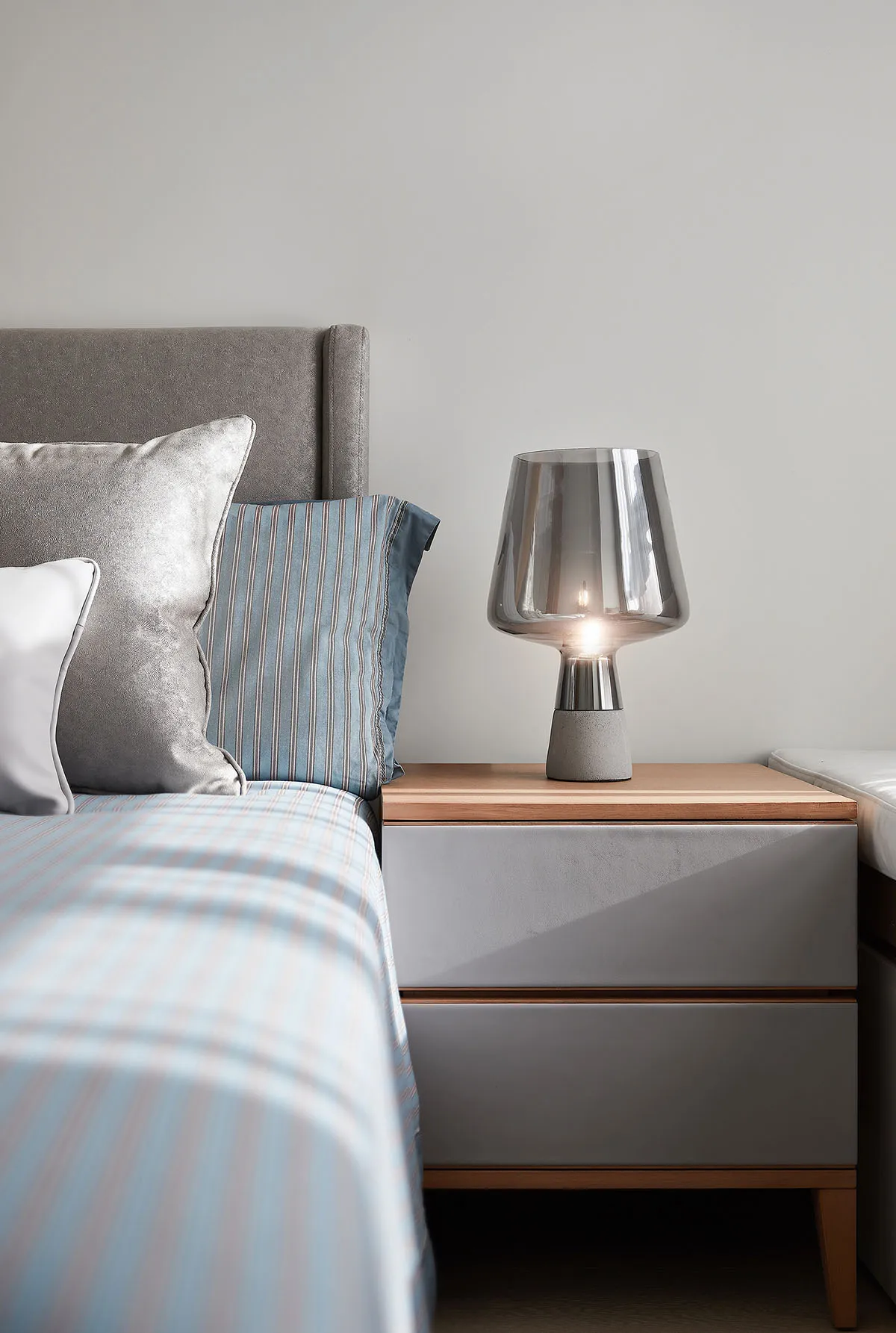 A modern bedroom featuring a wooden headboard, concrete bedside table, and glass lamp.