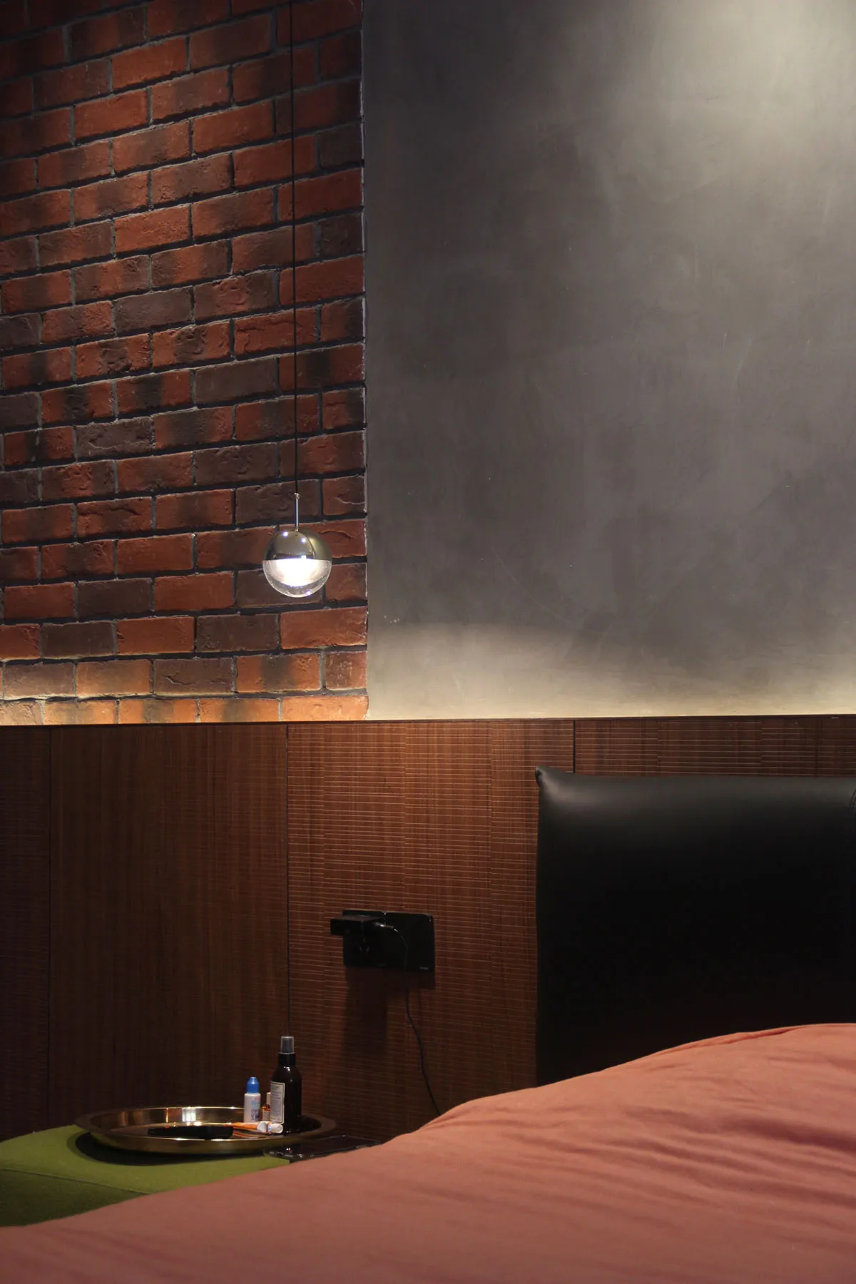 A modern bedroom with brick accent wall, wooden panels, a leather headboard, and metallic bedside lamp.