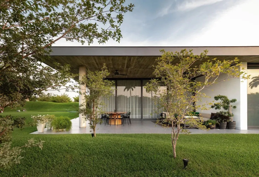 A modern house with a concrete exterior has a large open-air patio with a dining table and chairs surrounded by greenery and 