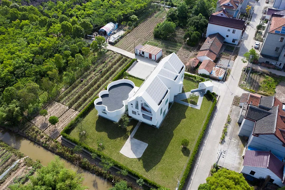 A modern house with a white, concrete exterior surrounded by greenery and a small pool.