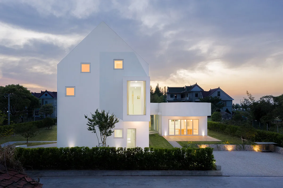 A modern white house with concrete exterior at dusk with illuminated rooms.
