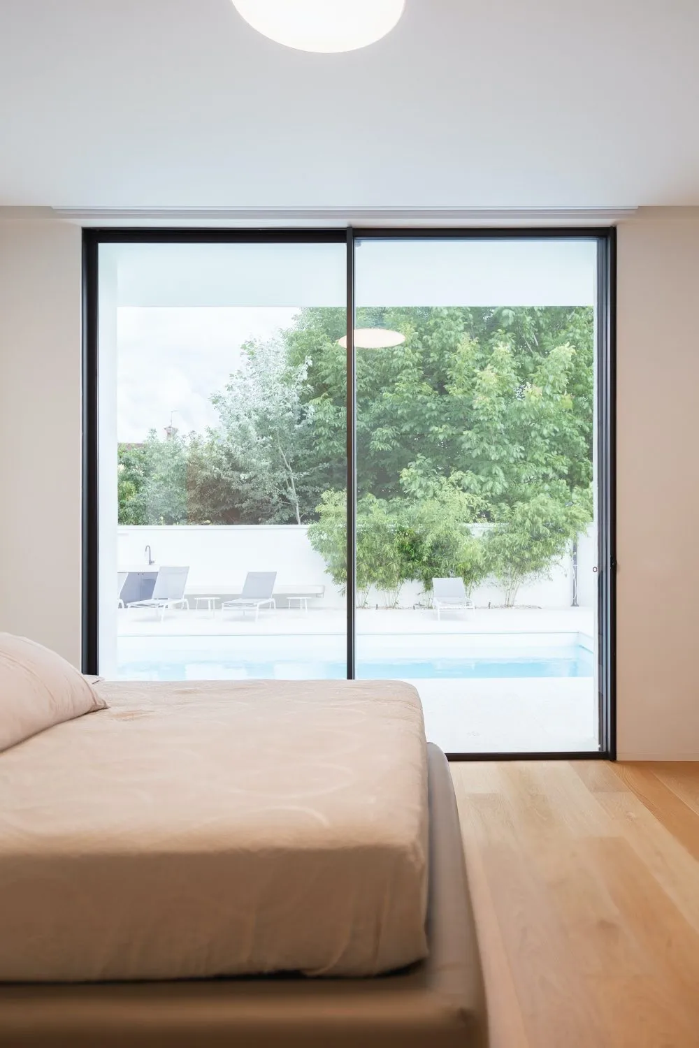 Modern minimalist bedroom with a large glass window overlooking a pool and garden, featuring wooden flooring and a comfortabl