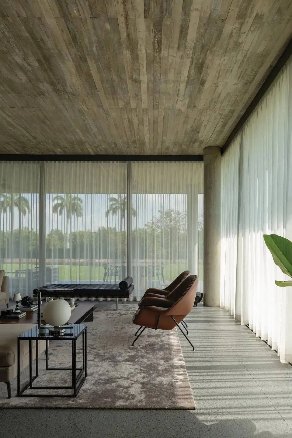 A minimalist living room with concrete ceiling and floor, featuring large windows, a brown armchair, a dark coffee table, and