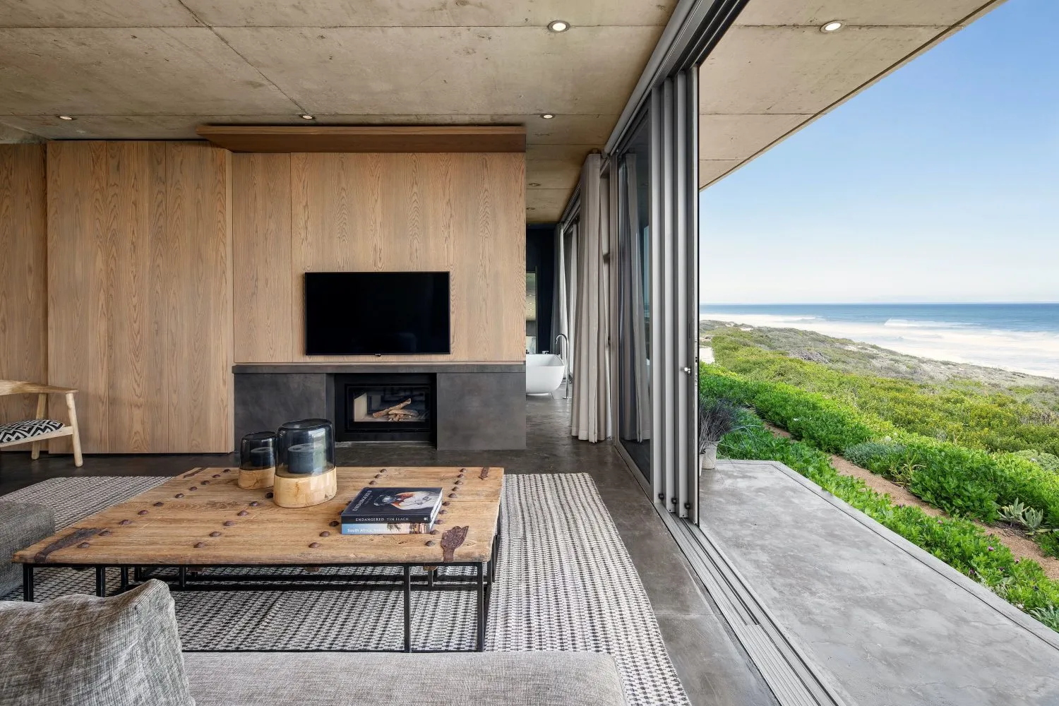 A modern living room with wooden furniture, large windows overlooking the sea, and a fireplace.