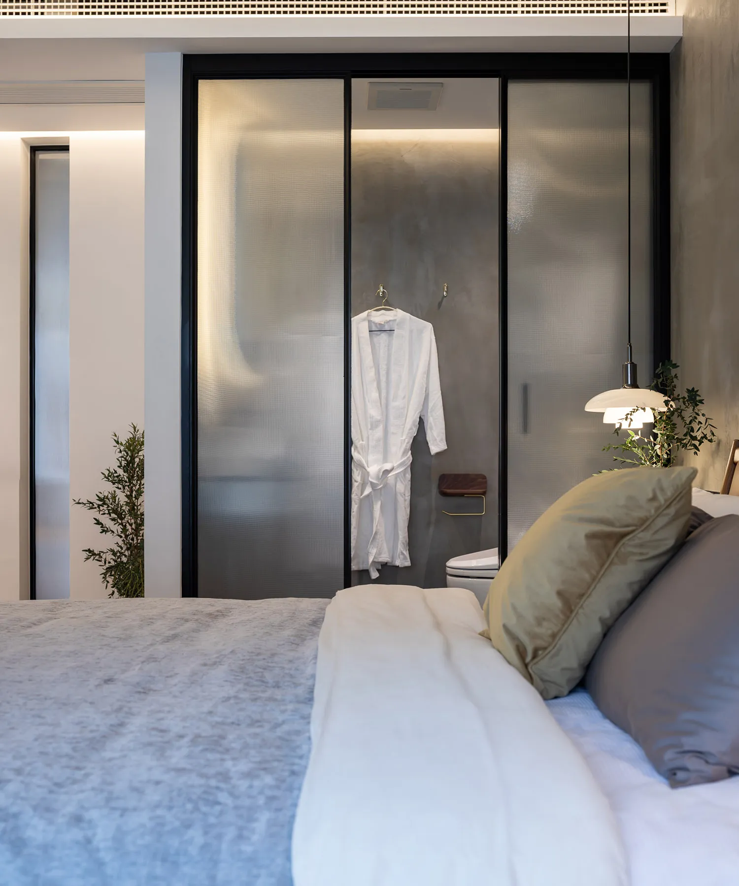Bedroom featuring a white bed with a beige pillow, a glass sliding door with a white robe, and a plant in the corner.