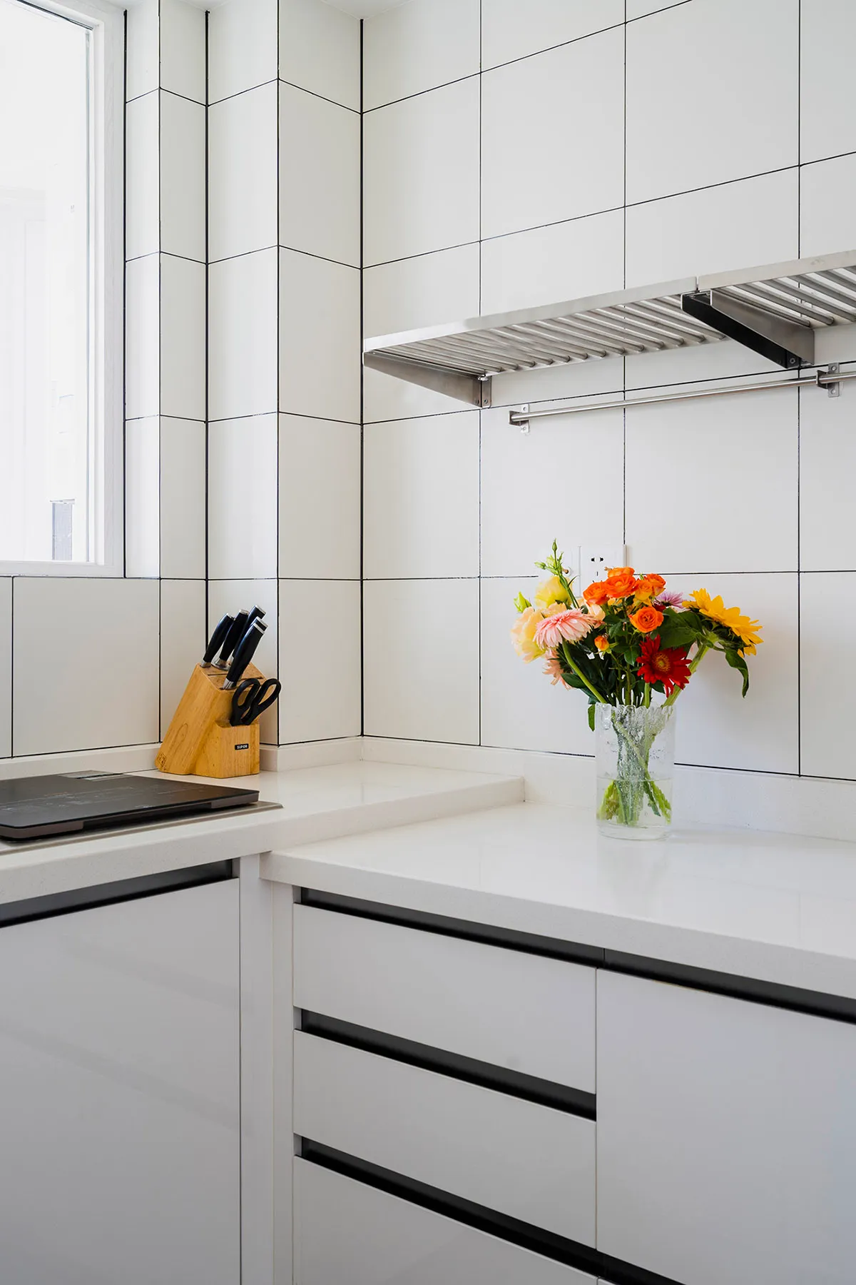 A minimalist kitchen with white ceramic tiles and black trim features a vase of colorful flowers on the countertop.