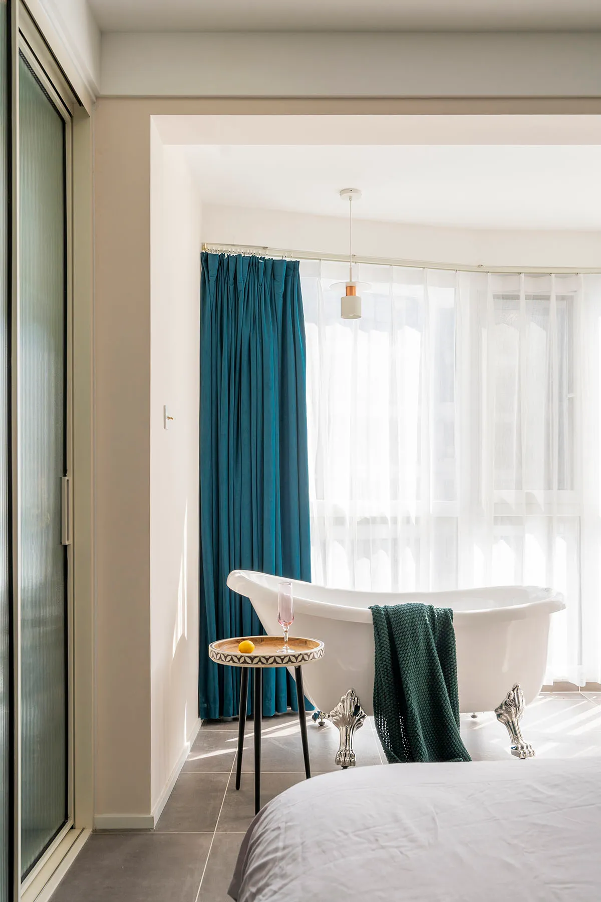 A modern bedroom featuring a white bathtub with ornate legs, blue velvet curtains, and a white bedspread, with a small side t