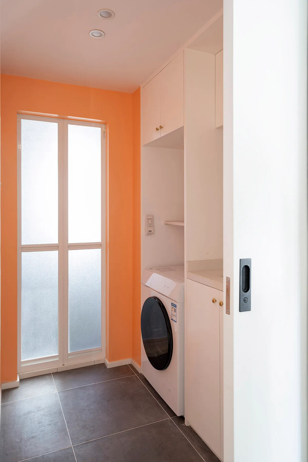 Laundry room with white appliances, orange walls, frosted glass door, and concrete tile flooring.