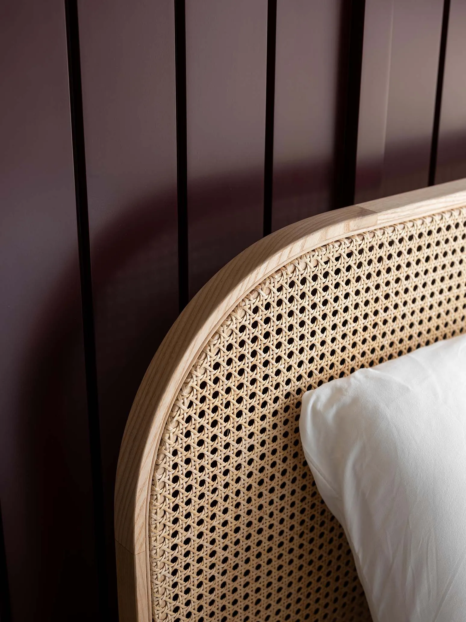 A modern bedframe with a woven cane headboard and a white pillow against a dark brown wooden wall.