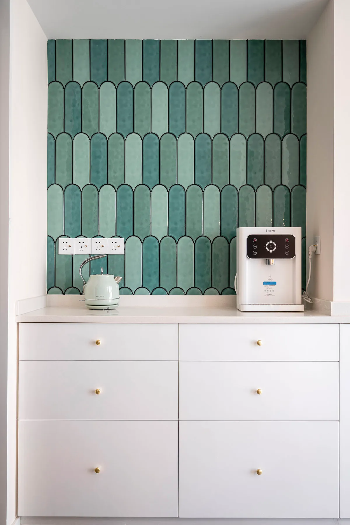 A modern kitchen with white cabinets, green ceramic tiles, and a white countertop.