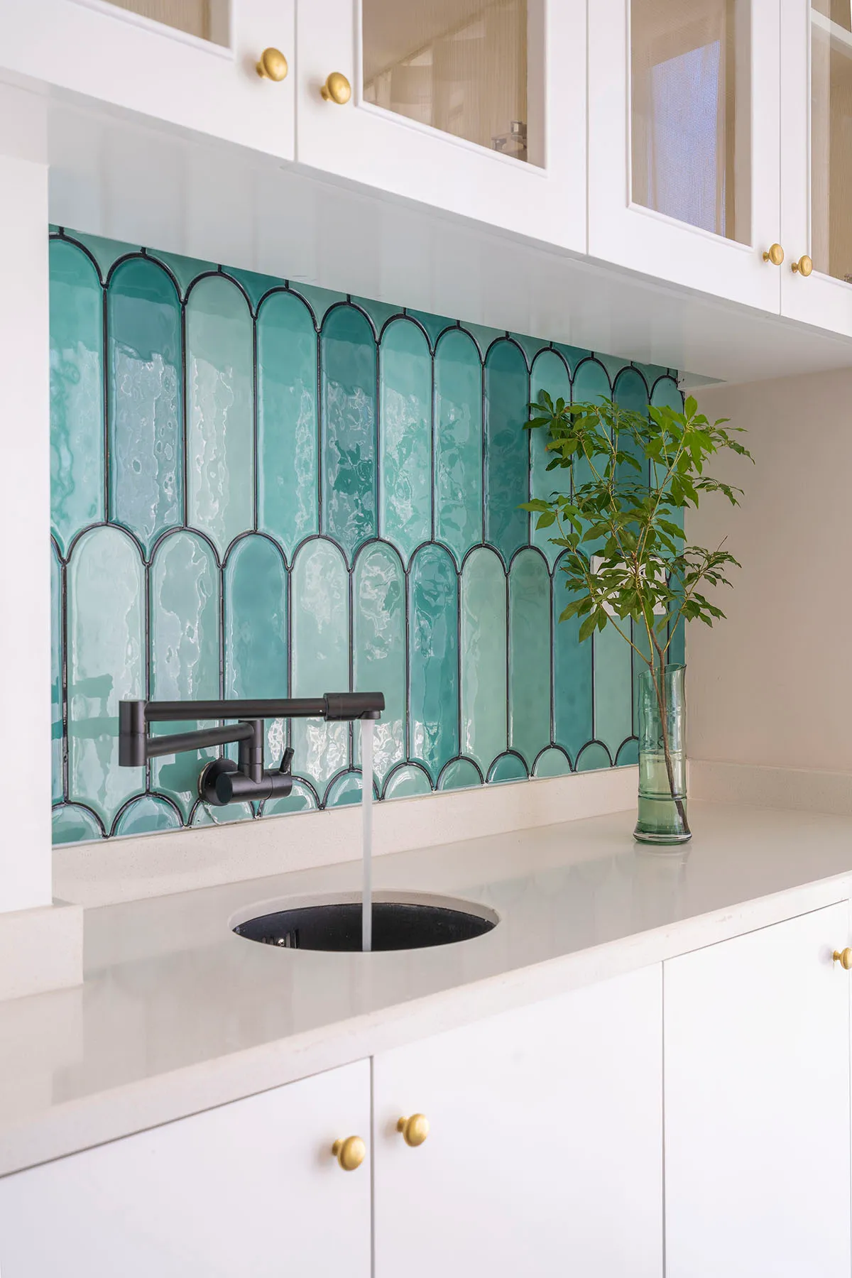 Modern kitchen with white cabinetry, glass tiles, and black faucet under white cabinets with glass-front doors.