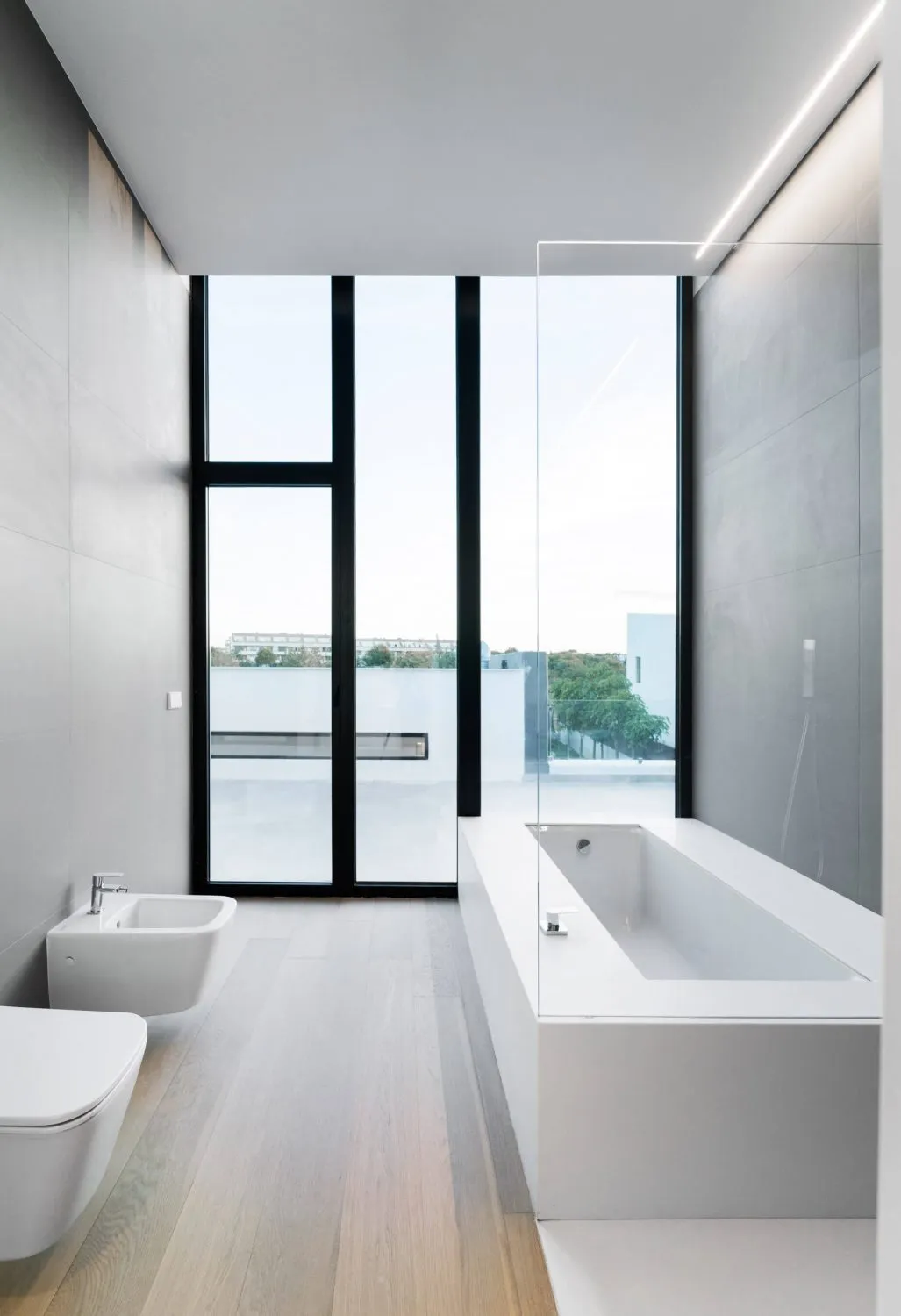 A modern bathroom with a minimalist design, featuring a concrete bathtub, a bidet, and a toilet with a light wooden floor.