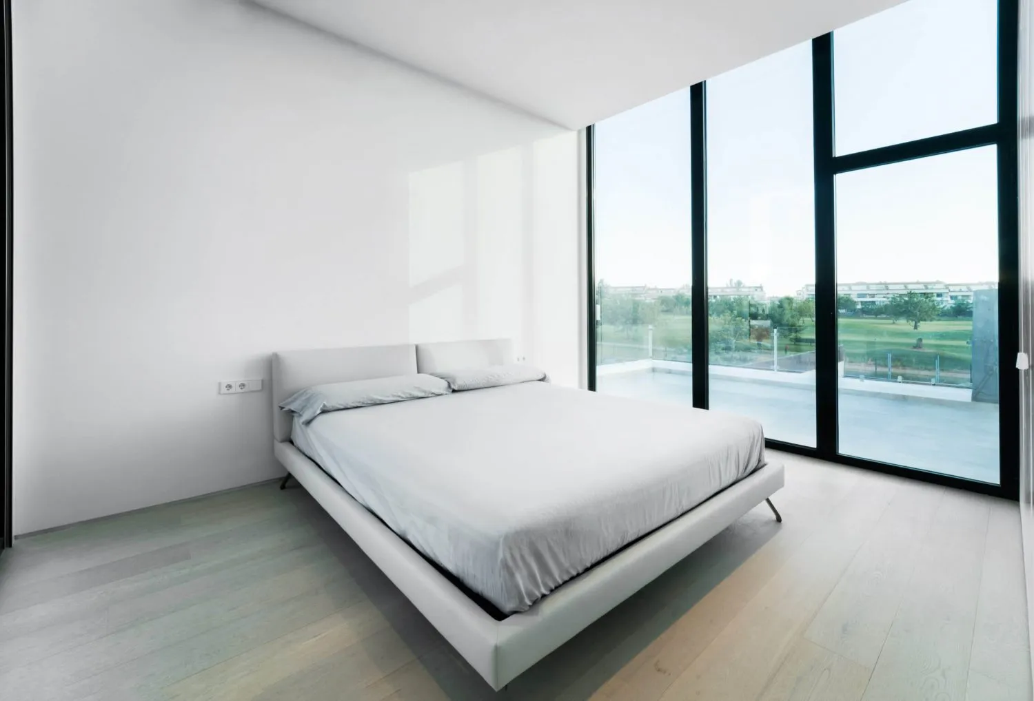 Modern minimalist bedroom with white furniture, glass windows, and wooden flooring.