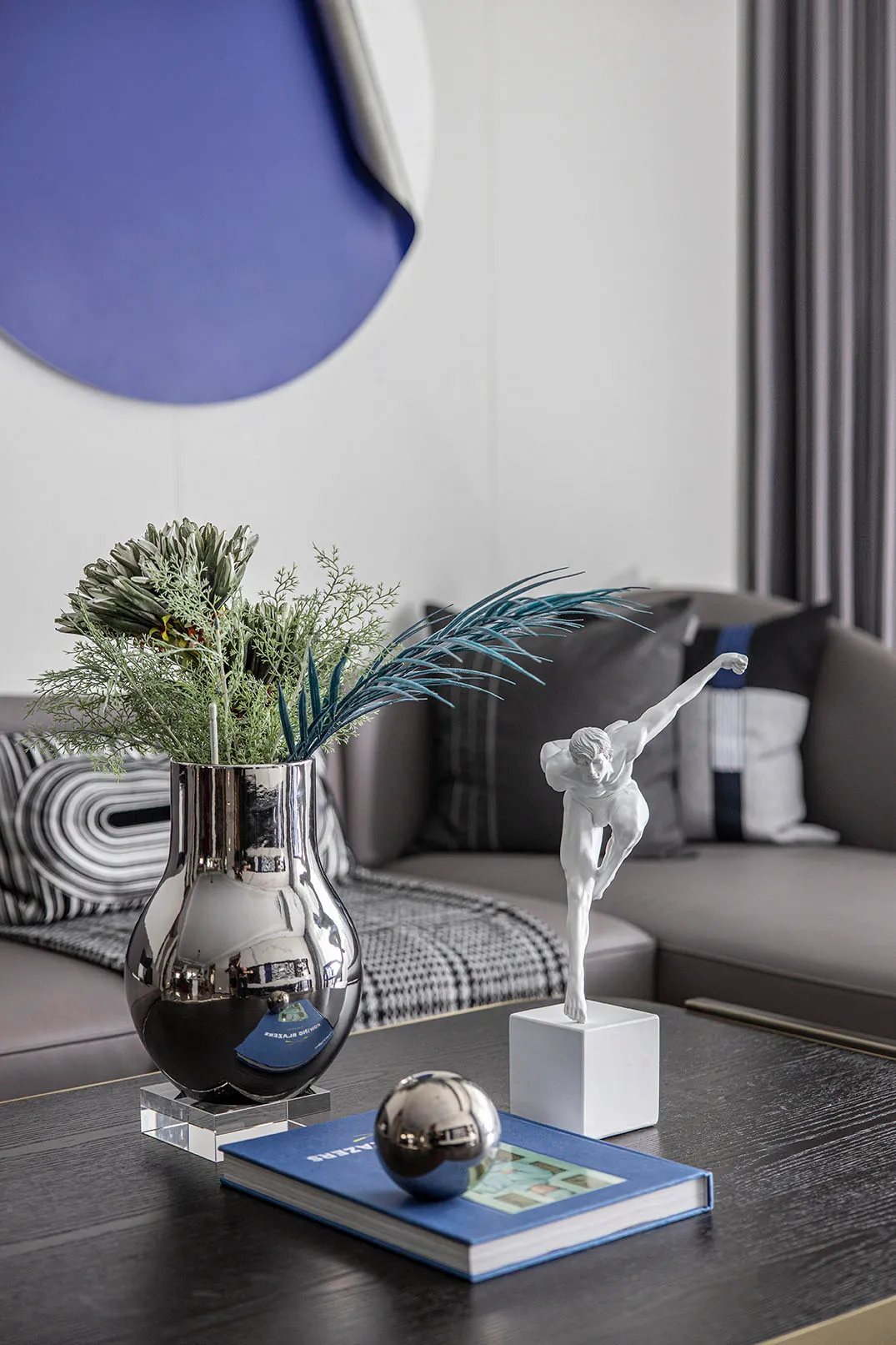A modern interior design featuring a metallic vase with greenery, a porcelain statue of a dancer, an acrylic ball, and a book