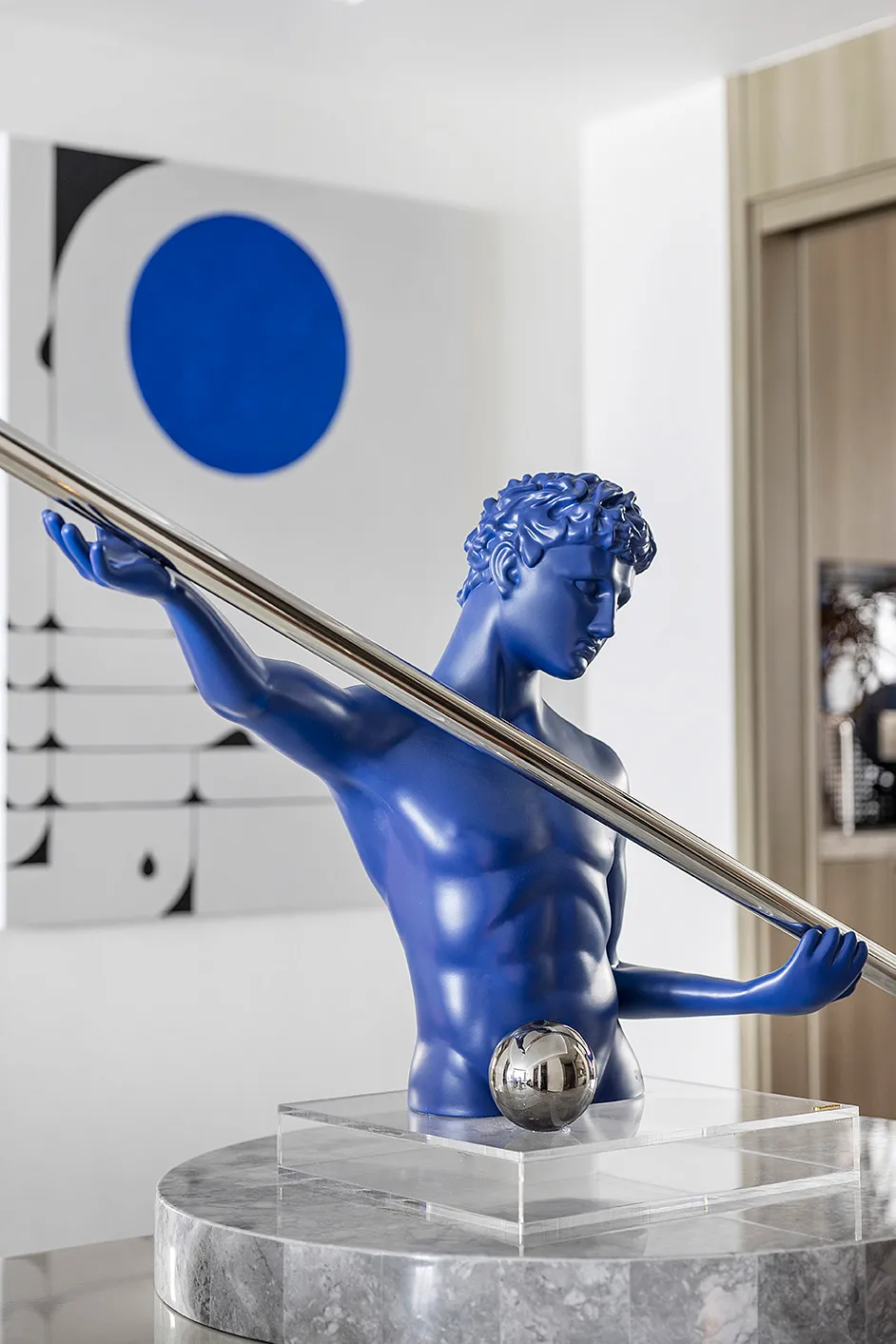 A blue sculpture with a metallic shaft and ball base, positioned on a marble pedestal.