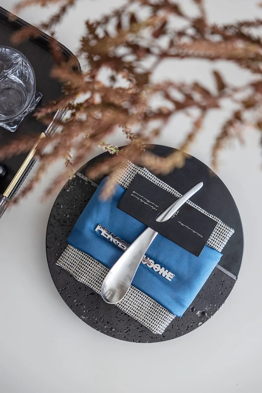 Modern dining table setting with a textured black stoneware plate, a blue textile napkin adorned with metallic letters spelli