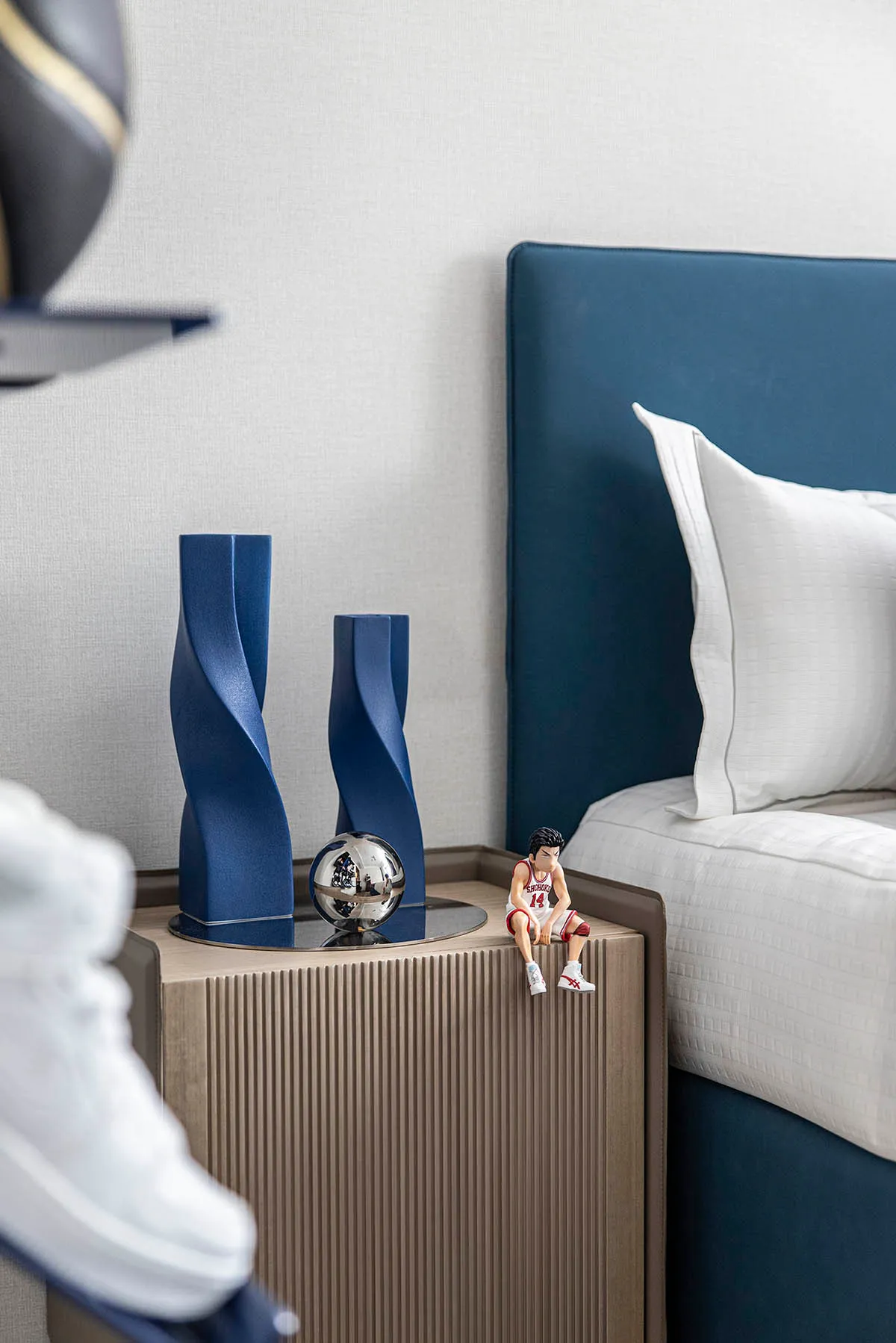 A modern bedroom with a blue upholstered headboard, white bedding, and a wooden nightstand with blue cylindrical decorations