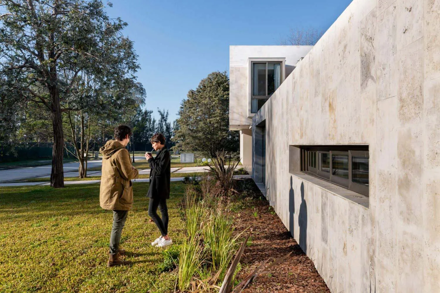 A modern house with a concrete exterior featuring a large window and greenery around it.