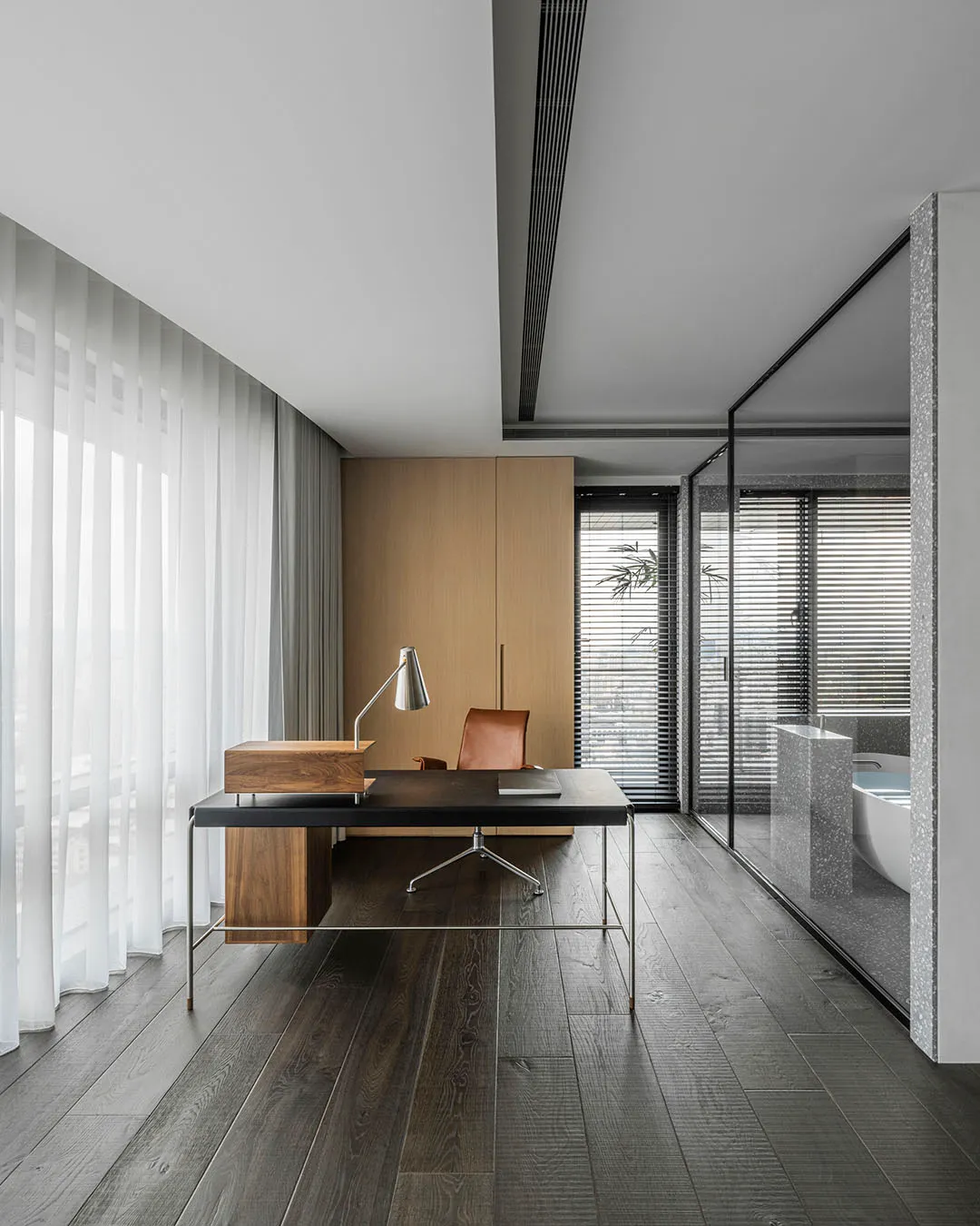 Modern office space with a wooden desk, leather chair, and lamp, featuring a glass-walled bathroom and large windows with she