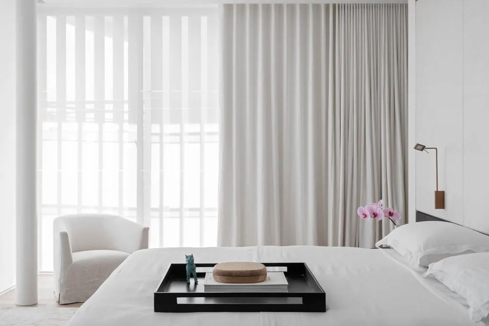 A minimalist bedroom with a white bed, white curtains, and a black tray with decorative items.