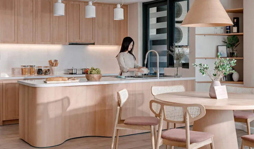 A modern kitchen with light-colored wood cabinets and a matching breakfast bar, featuring a woman washing dishes at the sink.