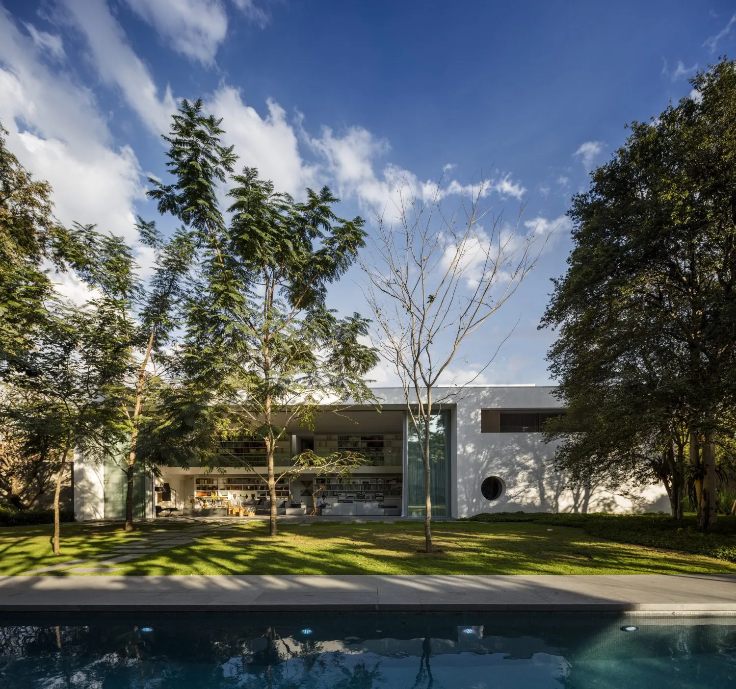 A modern house with white stucco exterior surrounded by green trees and a swimming pool.
