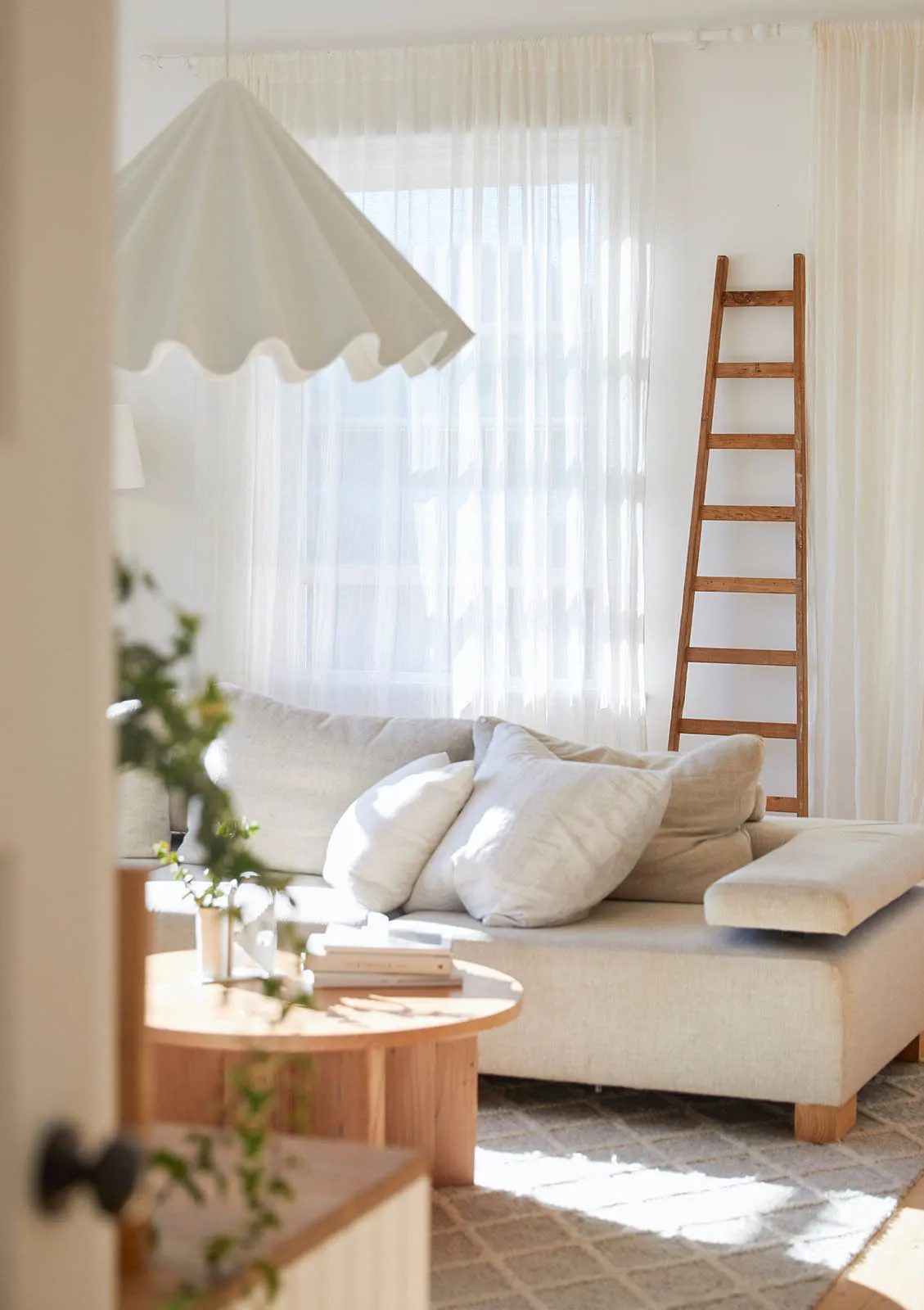 A white minimalist living room with a couch, a ladder, and a round wooden table with a plant.