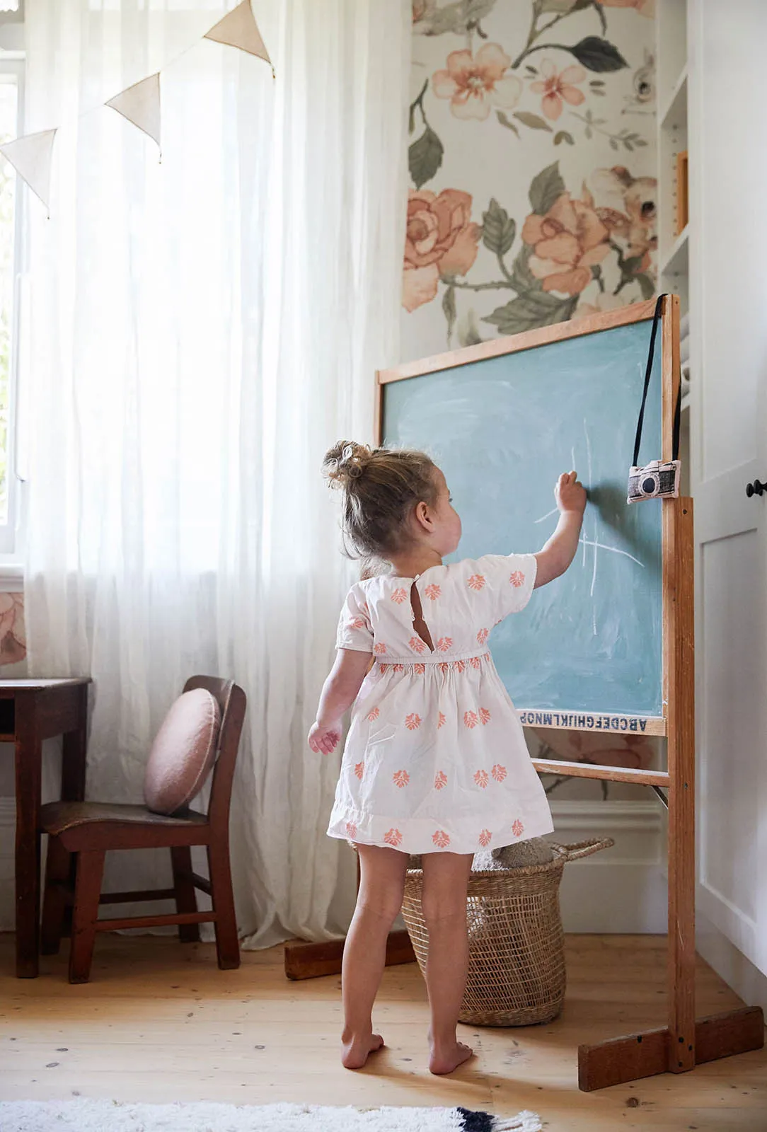 A young child stands at a wooden easel drawing on a chalkboard in a cozy, flower-patterned children's room.