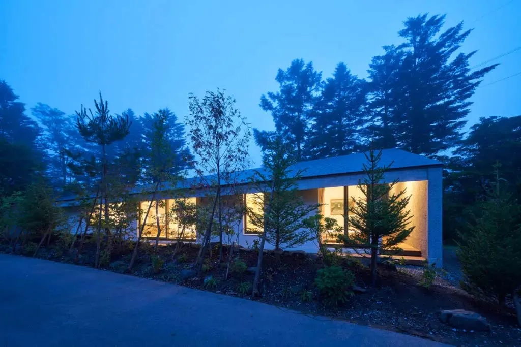 A modern residential house with illuminated windows and external lights, surrounded by greenery and tall trees.