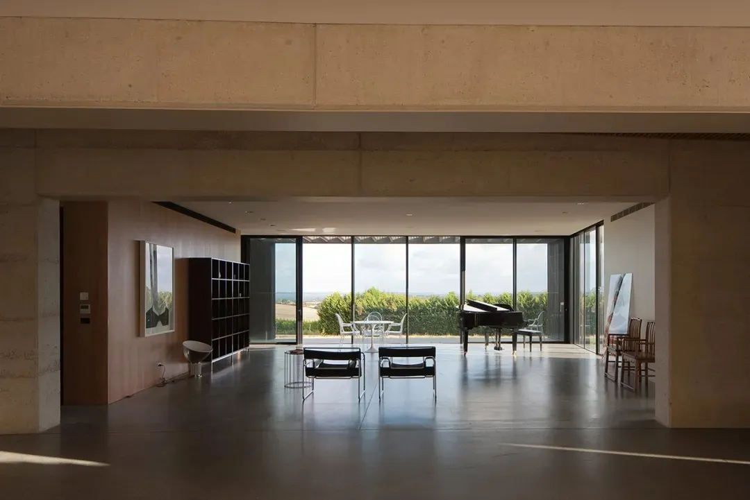 A modern living room with concrete flooring and walls, featuring large windows and minimalist furniture.