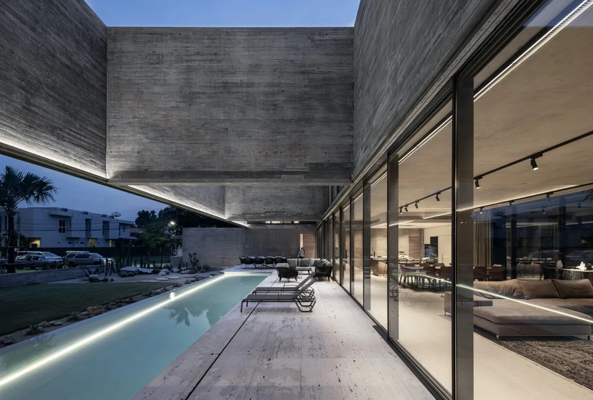 A modern, contemporary house with a concrete facade, featuring a pool and outdoor seating area, and an illuminated interior w