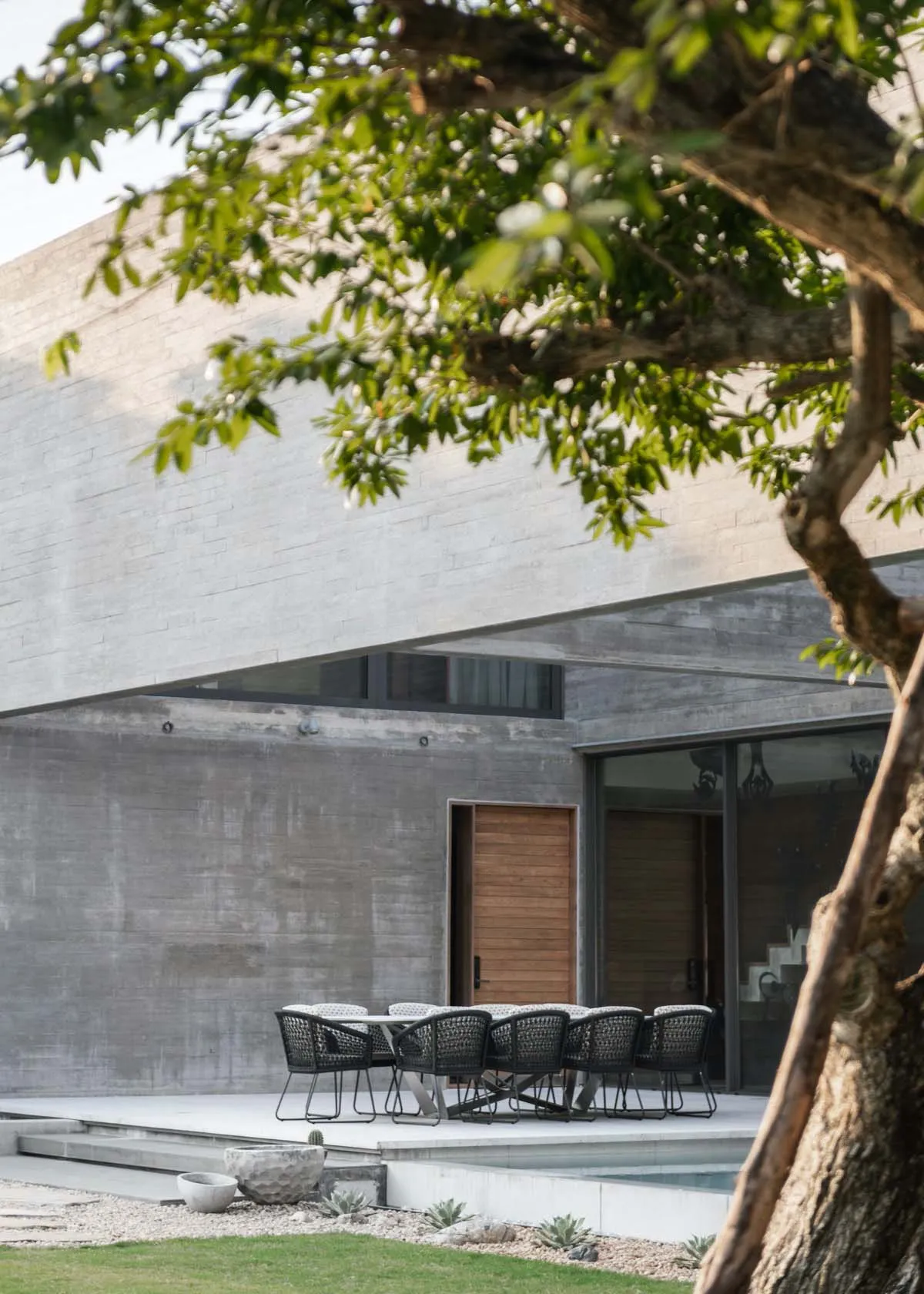A modern minimalist outdoor dining area with a concrete wall and a tree in the foreground.