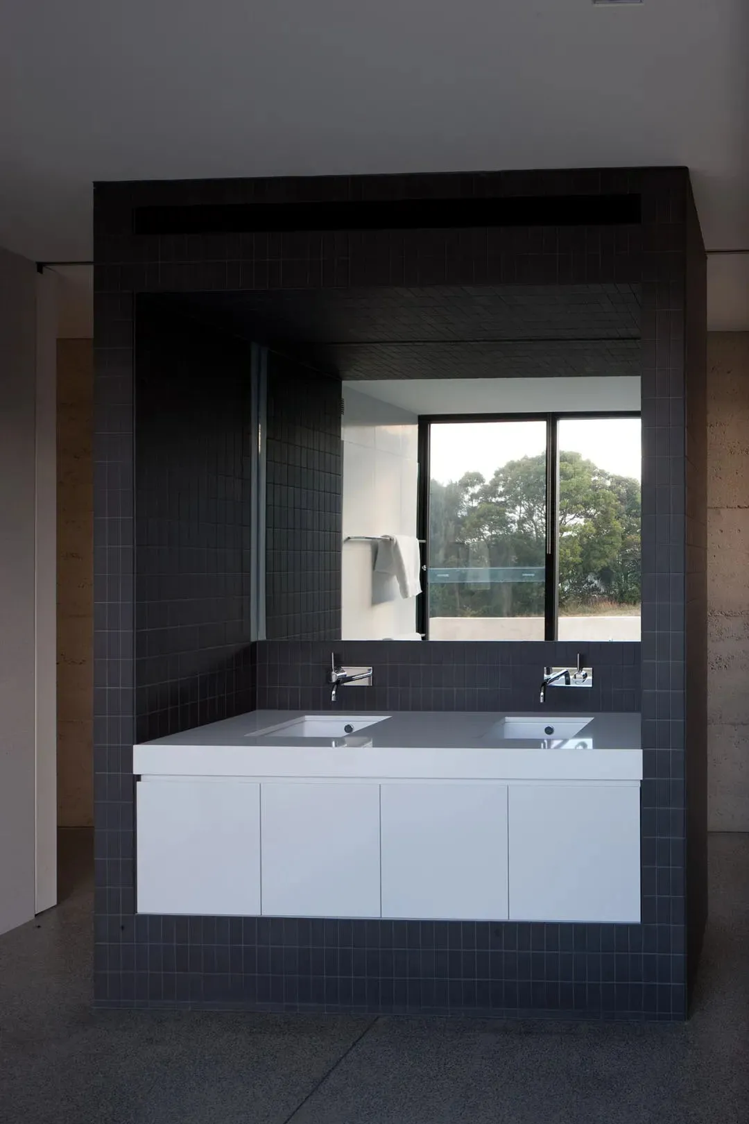 A modern residential bathroom with dark tiled walls, glass shower doors, and a concrete countertop.