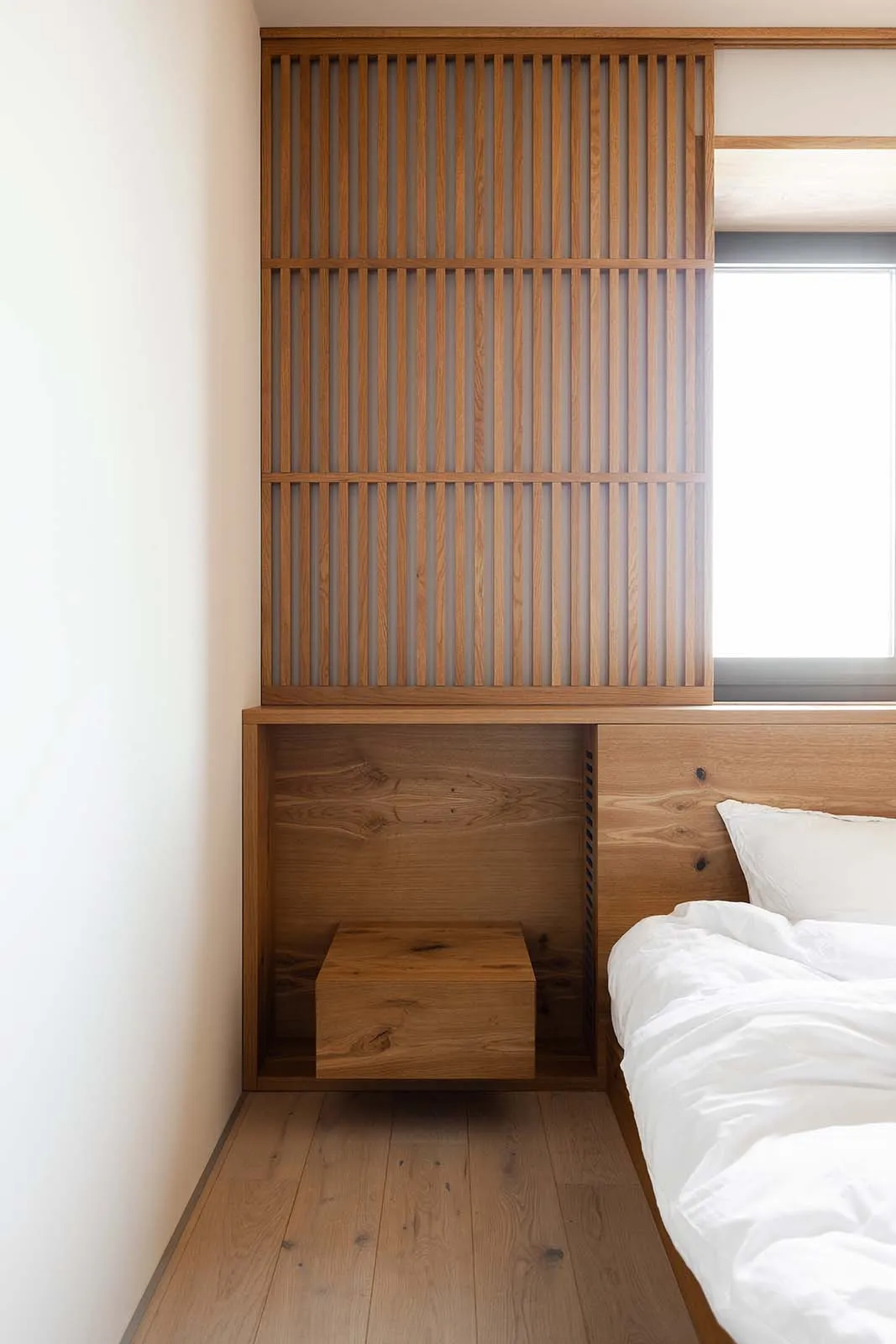 A modern, minimalist bedroom with wooden storage units, white bedding, and a window.