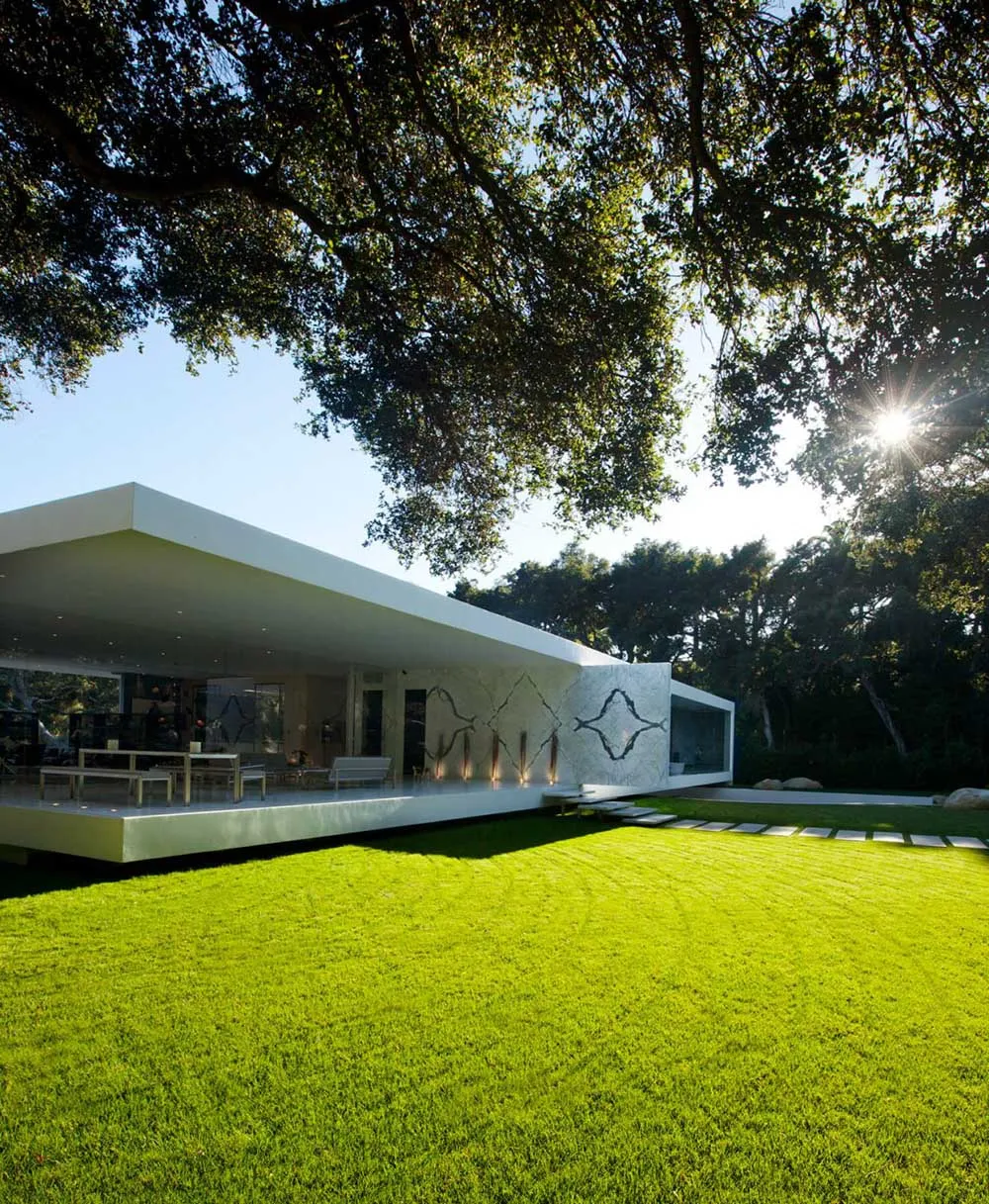 Modern white house with a concrete exterior and large glass windows, surrounded by green grass and trees.