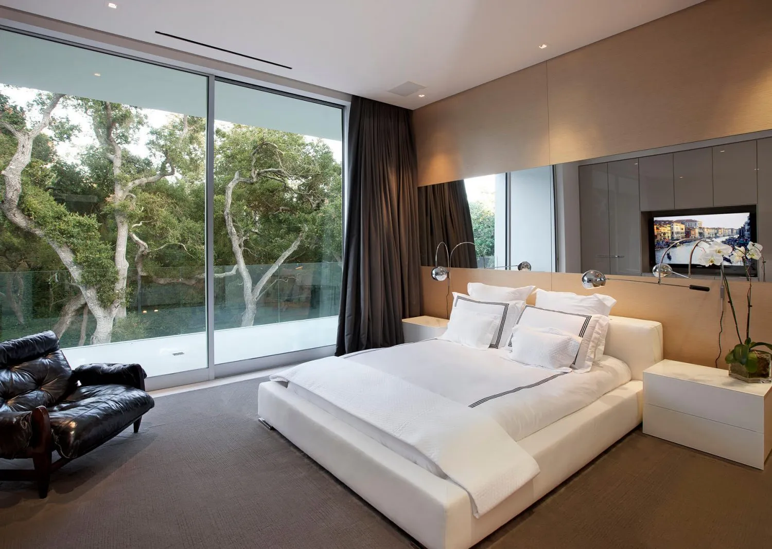 Modern residential bedroom featuring textured wood and leather furniture, large windows overlooking trees, and a sleek, minim