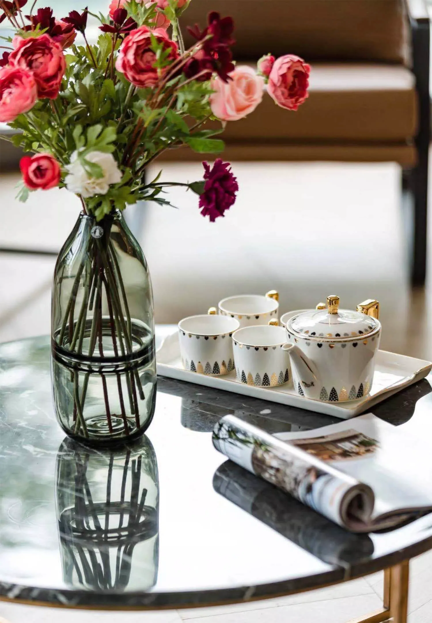 A modern interior featuring a glass coffee table with a vase of vibrant flowers, a teapot and cups, and a rolled-up magazine.