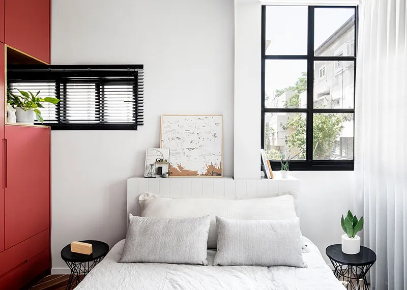 A minimalist bedroom featuring a white bed with grey pillows, a red cabinet, and a window with black frame.