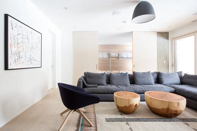 Modern minimalist living room featuring a large sectional sofa, wooden coffee tables, concrete floor, and abstract artwork.