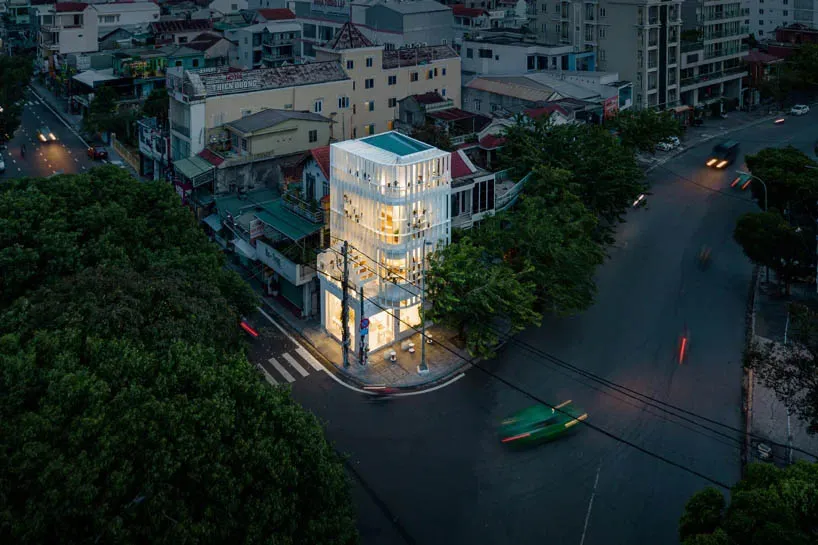 A modern glass building exterior at dusk with illuminated windows and a green car on the street.