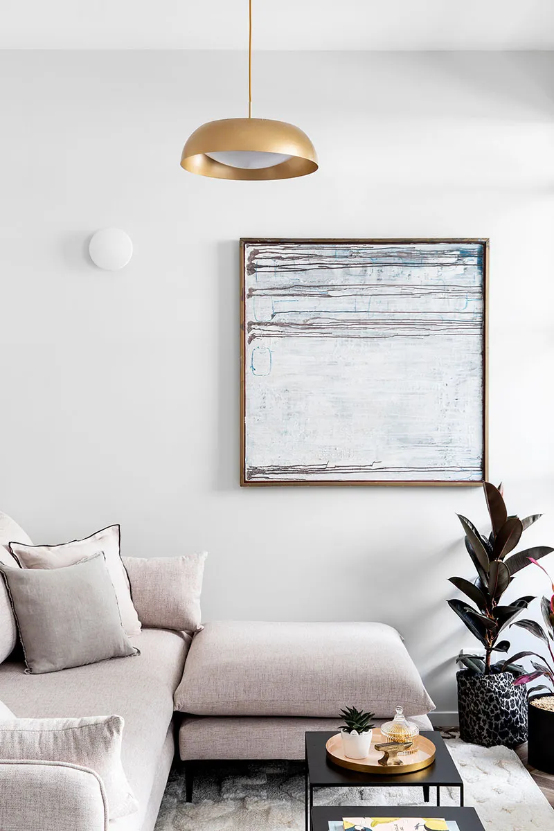 A modern living room with a beige sofa, pendant light, abstract art, and a small coffee table with a plant and candle.