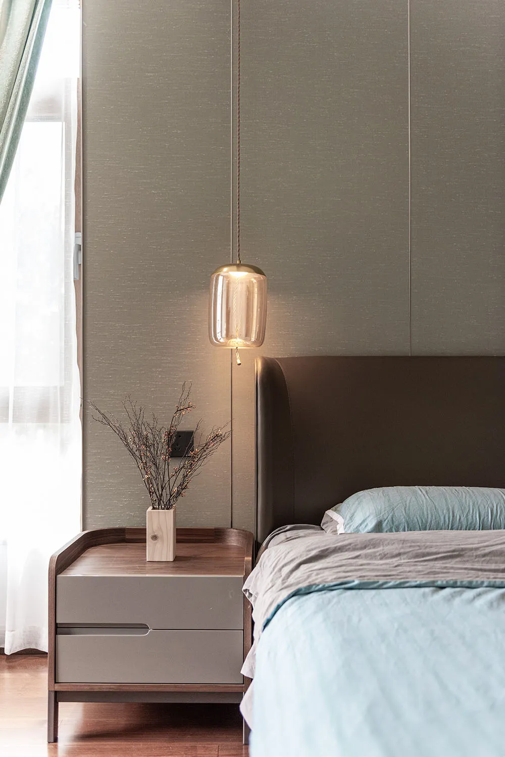 Modern minimalist bedroom with light blue bedding, wooden nightstand, and green foliage on the nightstand.