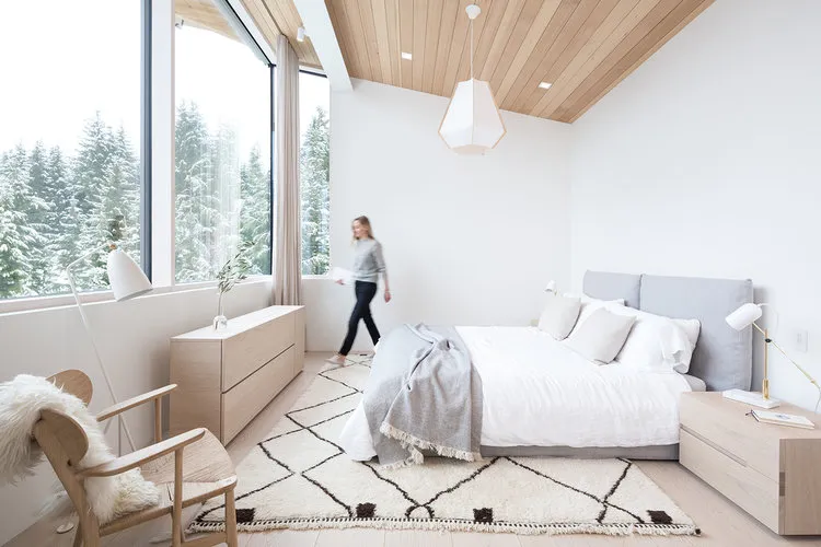 A modern minimalist bedroom with white walls, wooden furniture, and a large window overlooking snowy trees.