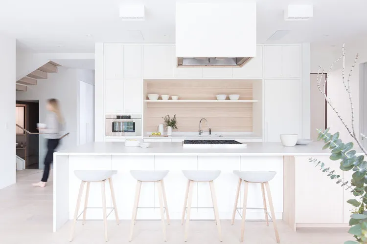 A modern minimalist kitchen featuring a white island with four stools, light-colored wood cabinets, and stainless steel appli