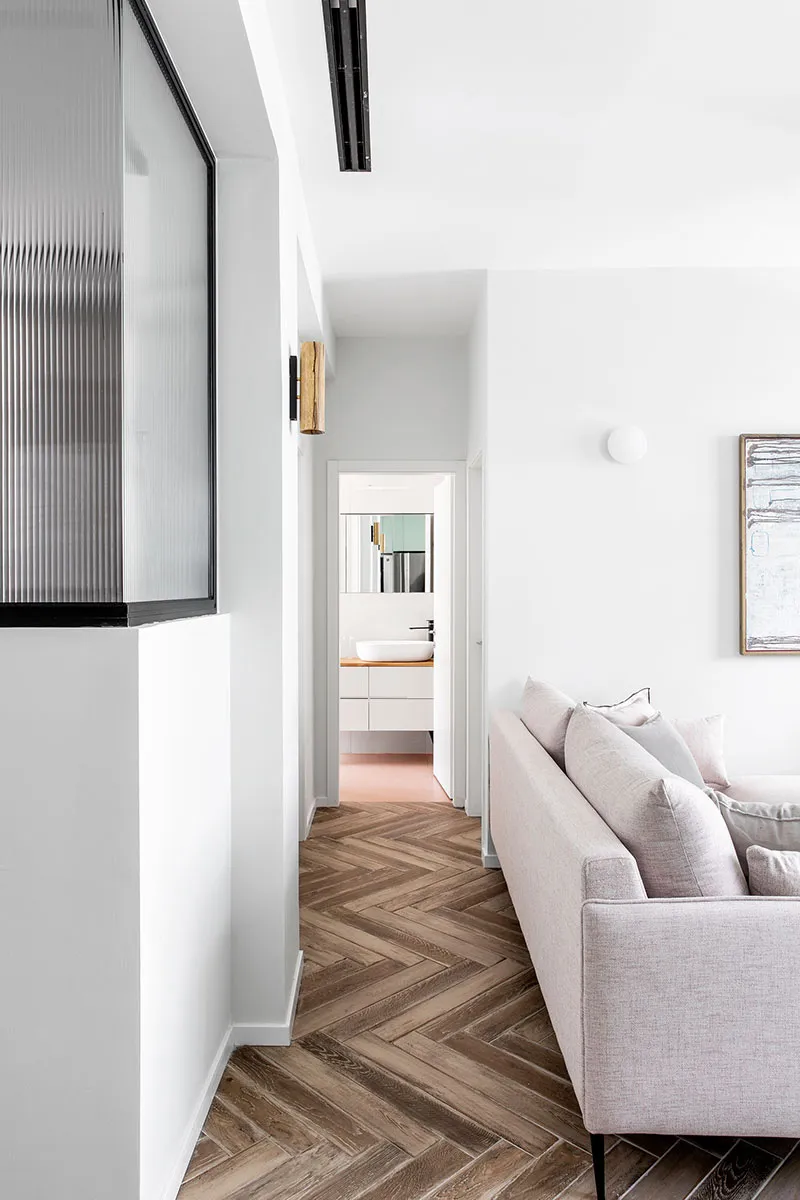 A modern minimalist living room with a beige couch, wooden herringbone floor, and a bathroom in the background.